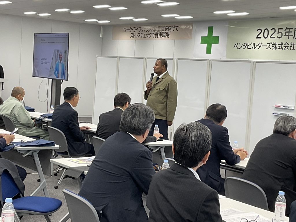 安全衛生大会の舞台上で、「個人のちがいを、組織の力へ」をテーマに講演するにしゃんたの写真。安全大会講師としての実績が分かる1枚。